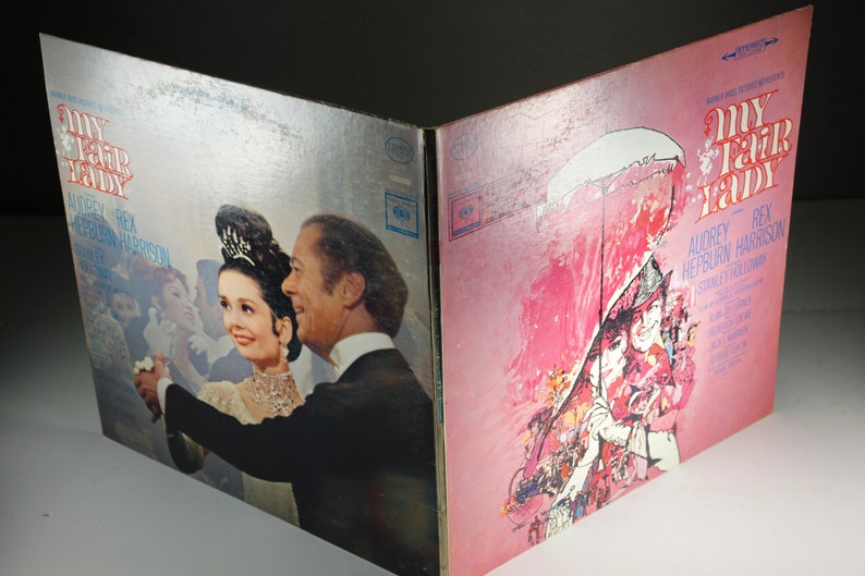 May include: A vintage vinyl record album cover for the musical "My Fair Lady" starring Audrey Hepburn and Rex Harrison. The front cover features a colorful illustration of the characters in a scene from the musical. The back cover features a photo of Audrey Hepburn and Rex Harrison dancing.