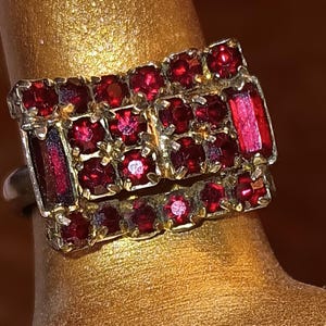 May include: A rectangular ring featuring a cluster of small, round, deep red faceted stones and two larger, elongated, rectangular red stones set in a metallic frame. The design is arranged in a grid-like pattern with multiple prongs securing each stone.