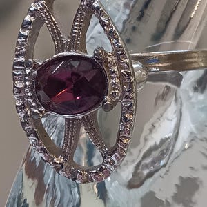 May include: Silver-toned ring featuring an oval, deep red gemstone. The ring has an openwork design with decorative detailing around the gemstone and along the band. The ring is set against a blurred background.