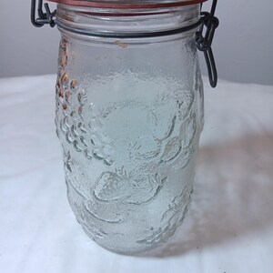 Puede incluir: Un tarro de cristal transparente con tapa abisagrada y junta de goma roja. El tarro tiene una superficie texturizada con un diseño de fruta. El cierre metálico es negro. El tarro está vacío y sobre una superficie blanca.