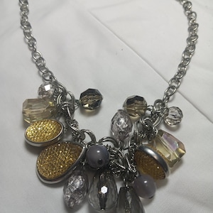 Puede incluir: Collar plateado con una variedad de colgantes. Los colgantes incluyen cuentas en forma de lágrima, facetadas y redondas en tonos dorados, plateados y grises. El collar tiene un diseño de eslabones de cadena.