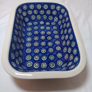 May include: Rectangular ceramic baking dish with a white rim and a blue interior. The interior is decorated with a pattern of green, red, and white circles. The dish has a glossy finish and is likely used for cooking or serving food.