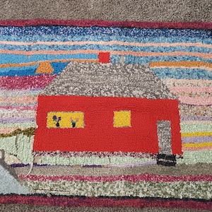 Puede incluir: Alfombra hecha a mano con un paisaje vibrante. Una casa roja con techo gris y ventanas amarillas es el punto focal. El fondo representa un cielo colorido y un árbol verde. La alfombra tiene un borde rojo oscuro.