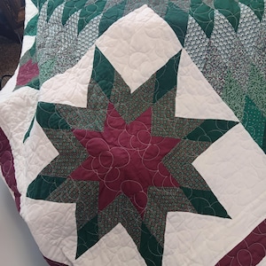 Può includere: Una trapunta fatta a mano con un grande disegno a stella al centro. La stella è composta da tessuto bordeaux, verde e fantasia, su uno sfondo bianco. La trapunta presenta una varietà di motivi geometrici e colori, tra cui verde, bordeaux e bianco.