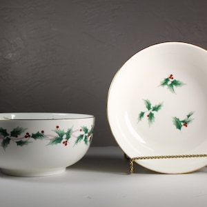 May include: Two white ceramic bowls with a gold rim and a holly leaf design. The smaller bowl has a single holly leaf design and the larger bowl has a repeating holly leaf design.