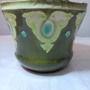 May include: A green ceramic planter with a flared shape. The planter has a glossy finish and is decorated with light green and turquoise floral designs. The rim has a textured, scalloped edge.