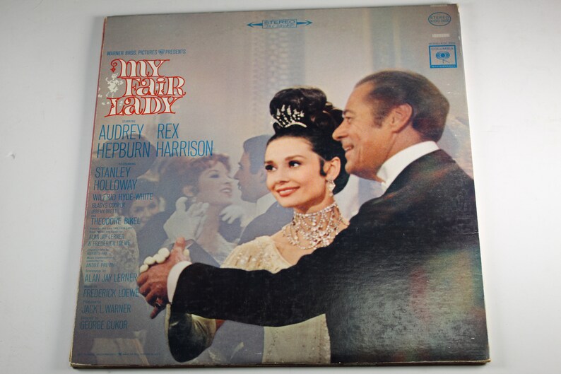 May include: A vintage vinyl record cover for the musical "My Fair Lady" starring Audrey Hepburn and Rex Harrison. The cover features a photo of Hepburn and Harrison in a ballroom scene.