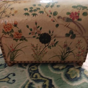 May include: A decorative chest with a floral and bird design. The chest is beige with a painted pattern of flowers, birds, and foliage in shades of pink, orange, green, and brown. The chest has a dark metal clasp and is trimmed with small, round studs.