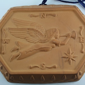 May include: A terracotta plaque with an embossed design of an angel blowing a trumpet. The plaque is octagonal and has a compass rose design in the lower right corner. The letters N, W, S, and E are visible around the compass rose.