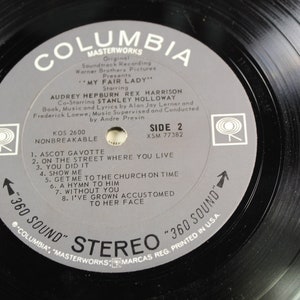 May include: A black vinyl record with a gray label. The label has the text "Columbia Masterworks" and "My Fair Lady" with the names of the actors Audrey Hepburn, Rex Harrison, and Stanley Holloway. The record is side 2 and has the song titles "Ascot Gavotte", "On the Street Where You Live", "You Did It", "Show Me", "Get Me to the Church on Time", "A Hymn to Him", "Without You", and "I've Grown Accustomed to Her Face".