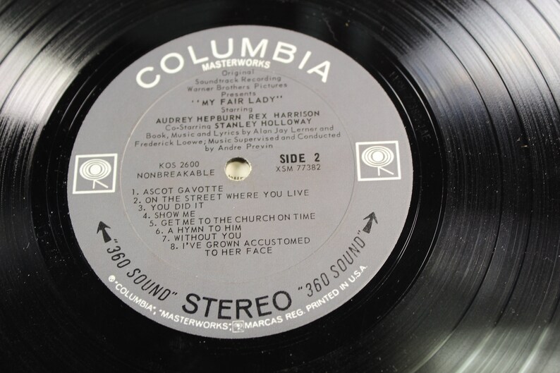 May include: A black vinyl record with a gray label. The label has the text "Columbia Masterworks" and "My Fair Lady" with the names of the actors Audrey Hepburn, Rex Harrison, and Stanley Holloway. The record is side 2 and has the song titles "Ascot Gavotte", "On the Street Where You Live", "You Did It", "Show Me", "Get Me to the Church on Time", "A Hymn to Him", "Without You", and "I've Grown Accustomed to Her Face".