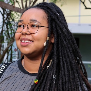 Op de afbeelding: Een jonge vrouw met lange zwarte dreadlocks, die een zwart-wit gestreept shirt, een zwarte bril en gouden oorbellen draagt. Ze glimlacht en kijkt naar de zijkant.