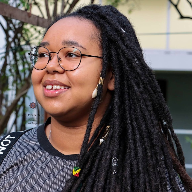 Op de afbeelding: Een jonge vrouw met lange zwarte dreadlocks, die een zwart-wit gestreept shirt, een zwarte bril en gouden oorbellen draagt. Ze glimlacht en kijkt naar de zijkant.