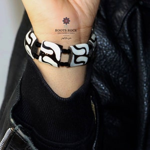 Könnte beinhalten: Ein Armband mit schwarz-weißem Muster wird am Handgelenk getragen. Das Armband besteht aus rechteckigen Perlen mit einem kontrastierenden schwarz-weißen Design. Der Markenname "ROOTS ROCK Afro Caribbean Design" ist sichtbar.
