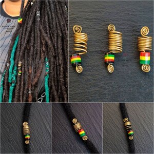 Op de afbeelding: Drie gouden metalen dreadlock kralen met een zwarte, groene en gele gestreepte kraal op elk. De kralen zijn op een zwarte achtergrond.