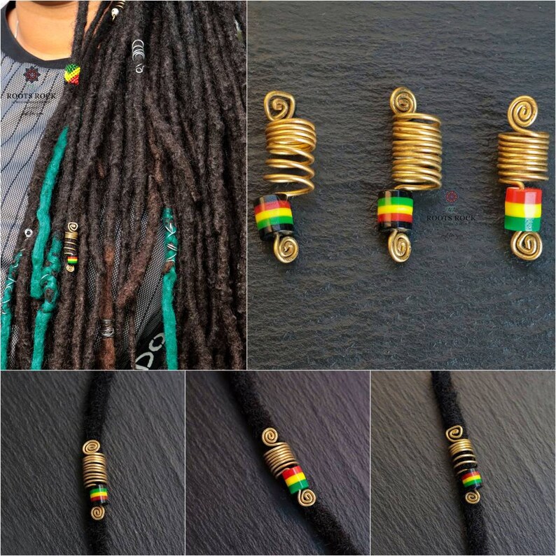 Op de afbeelding: Drie gouden metalen dreadlock kralen met een zwarte, groene en gele gestreepte kraal op elk. De kralen zijn op een zwarte achtergrond.