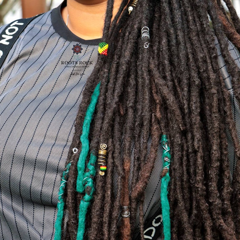 Op de afbeelding: Een persoon met lange, donkerbruine dreadlocks met groene en gouden kralen. De dreadlocks zijn gestyled met groene en blauwe touwtjes. De persoon draagt een grijs-wit gestreept shirt met de tekst "ROOTS ROCK" op de voorkant.