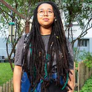 Op de afbeelding: Een persoon met lange, donkerbruine dreadlocks met groene en gele accenten. Ze draagt een grijs gestreept shirt met een zwarte band waarop "DO NOT" staat en een blauwe jeans.