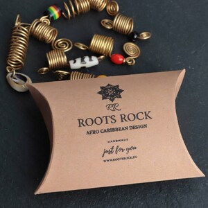 Op de afbeelding: Een bruine papieren zak met de tekst "Roots Rock Afro Caribbean Design" en "Handmade just for you" erop gedrukt. De zak bevat een set gouden haarspiralen.
