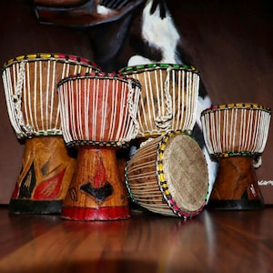 May include: Five wooden djembe drums with various designs and colours. The drums are arranged in a semi-circle on a wooden surface.
