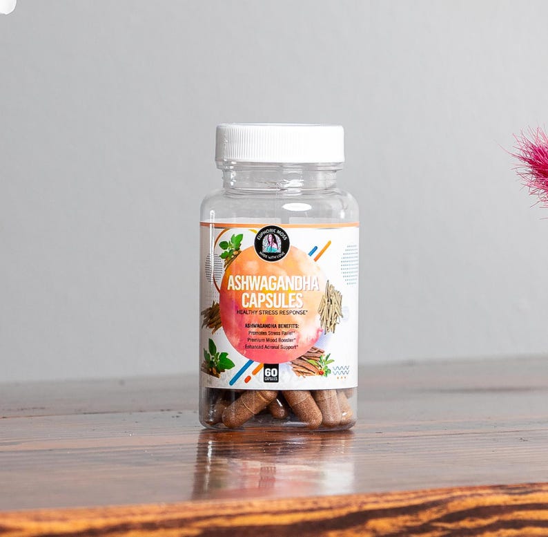 May include: A clear plastic bottle with a white lid filled with brown capsules. The label reads "Ashwagandha Capsules Healthy Stress Response" and lists benefits including "Promotes Stress Relief" and "Premium Mood Booster".