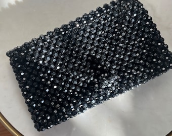 Op de afbeelding: Een zwarte clutch met kralen. De rechthoekige tas is bedekt met kleine, facetgeslepen zwarte kralen, wat een textuur geeft. De sluiting is verborgen. Geschikt voor avondkleding of speciale gelegenheden.