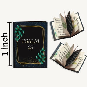 May include: A small, black book with a gold frame and green leaves. The book says "PSALM 23" on the front. The book is open to reveal the text "He maketh me to lie down in green pastures: he leadeth me beside the still waters."