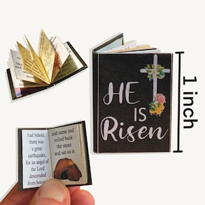 May include: A small, black book with the words "He is Risen" printed in white. The book is decorated with a white cross and flowers. The book is 1 inch tall.