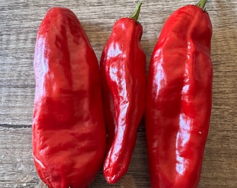 May include: Three vibrant red chili peppers arranged vertically. The peppers have a smooth, glossy skin with slight natural indentations and green stems at the top. Each pepper displays a distinct elongated shape with a rounded base and a tapered tip.