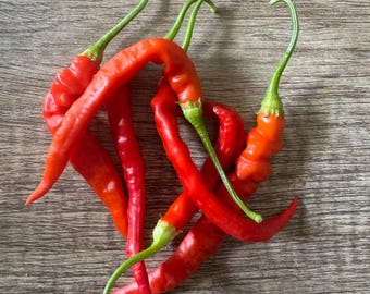 May include: A cluster of vibrant red chili peppers with green stems, arranged on a wooden surface. The peppers have a curved shape and a glossy texture, suggesting freshness. The image is a close-up shot, highlighting the details of the peppers.
