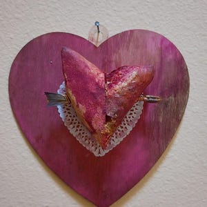 May include: A heart-shaped wooden wall hanging with a deep pink hue. The heart features a dried flower arrangement in shades of red and gold, mounted on a white doily. A small metal pin secures the piece to the wall.