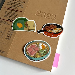 Könnte beinhalten: Drei illustrierte Aufkleber mit japanischem Essen. Der erste Aufkleber zeigt ein Sandwich mit einem Spiegelei, der zweite Aufkleber zeigt eine Pfanne mit Speck und Eiern, und der dritte Aufkleber zeigt eine Schüssel Ramen mit Schweinefleisch und einem Spiegelei.