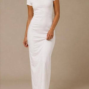 Puede incluir: Un vestido blanco largo hasta el suelo con cuello redondo y mangas cortas. La prenda presenta un cuerpo ajustado que se transforma en una falda recta de largo completo, confeccionada en un tejido sólido y opaco que llega al suelo.