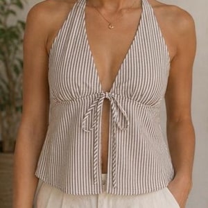 Pode incluir: Um top frente única sem mangas com um padrão de riscas verticais em cinzento-acastanhado e branco. A peça tem um decote em V profundo com um fecho de laço central no busto, criando um design frontal aberto até à bainha, com calções brancos.