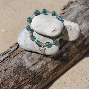 May include: A teal and silver beaded bracelet with a turtle charm. The bracelet is on a pile of gray stones and a piece of driftwood.