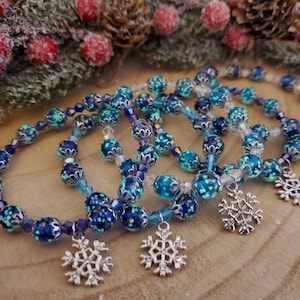 May include: Several beaded bracelets with blue and turquoise beads and silver snowflake charms. The bracelets are strung with clear beads and feature decorative silver accents. The bracelets are a winter holiday theme.