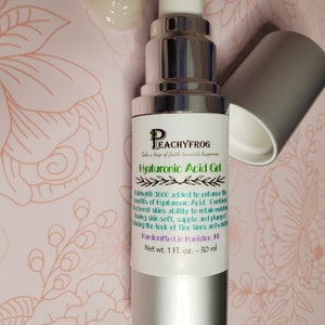 May include: A silver bottle of Hyaluronic Acid Gel with a white pump top. The label reads "Peachyfrog" and "Take a leap of faith towards happiness". The bottle is on a pink and green floral background. Net wt. 1 Fl. oz. - 30 ml.