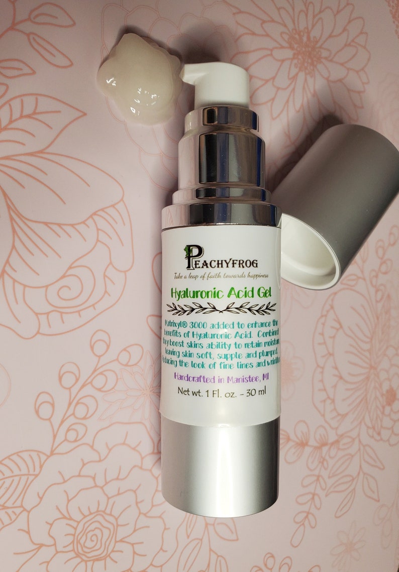 May include: A silver bottle of Hyaluronic Acid Gel with a white pump top. The label reads "Peachyfrog" and "Take a leap of faith towards happiness". The bottle is on a pink and green floral background. Net wt. 1 Fl. oz. - 30 ml.