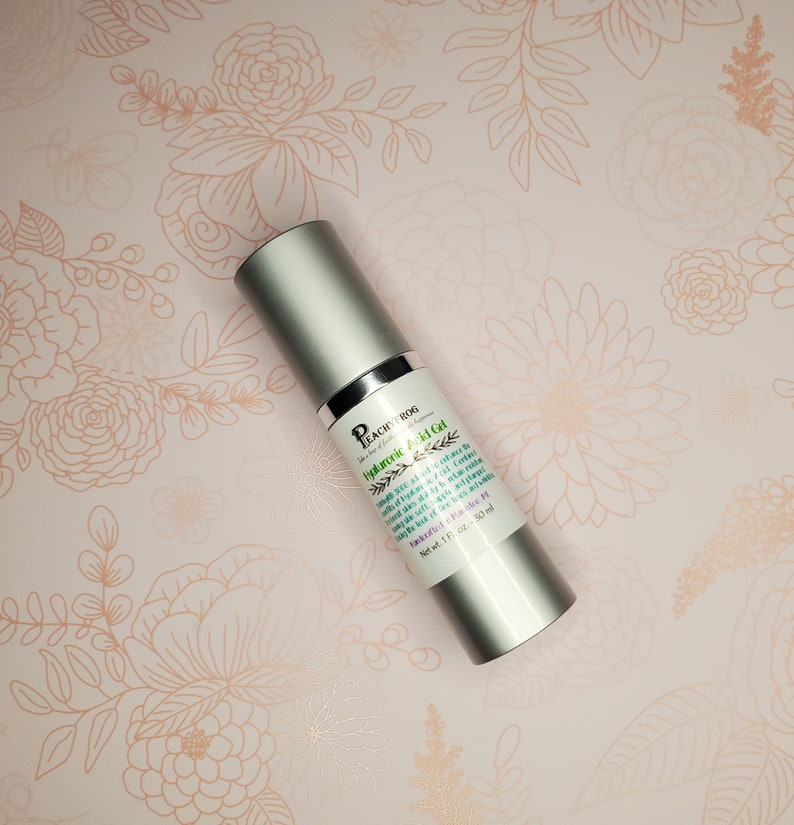 May include: A silver bottle of Peach & Bog "Hyduronic Acid Gel" skin care product. The bottle is labeled with the product name and ingredients. The bottle is on a pink background with a floral pattern.