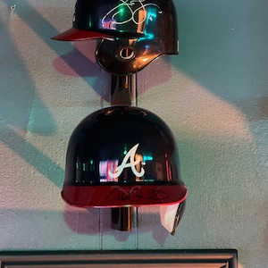 May include: Two baseball helmets hanging on a wall. The top helmet is black with a red brim and a white signature. The bottom helmet is black with a red brim and a white "A" on the front.