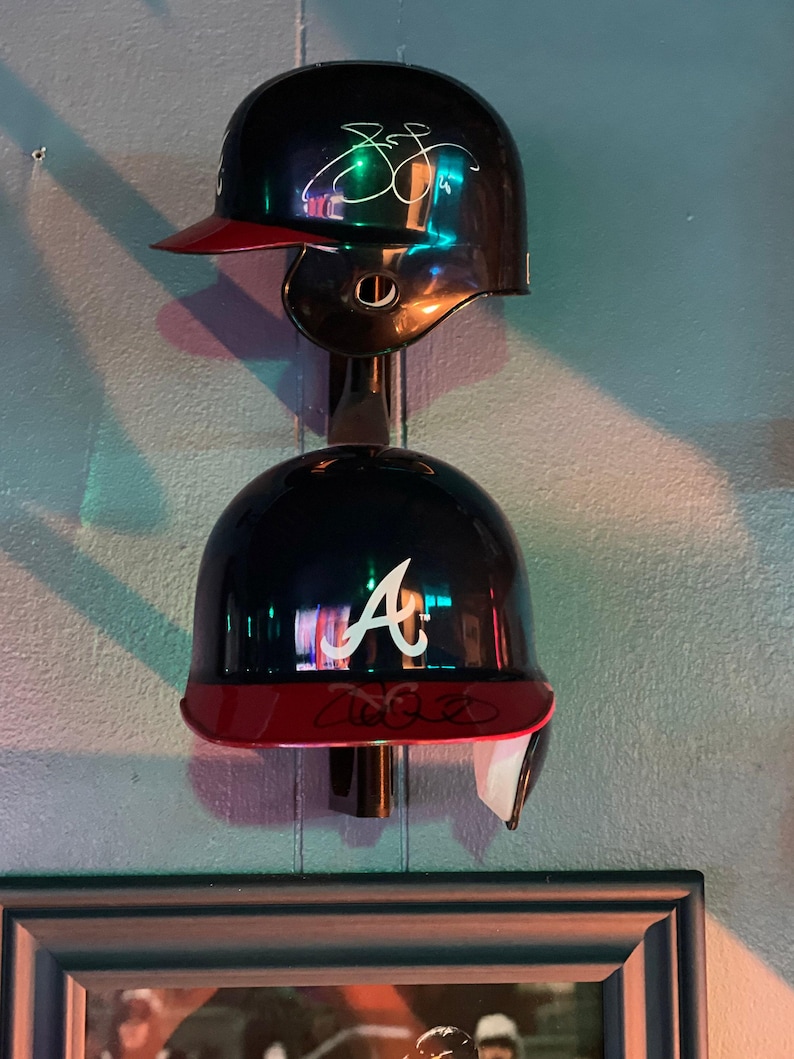 May include: Two baseball helmets hanging on a wall. The top helmet is black with a red brim and a white signature. The bottom helmet is black with a red brim and a white "A" on the front.