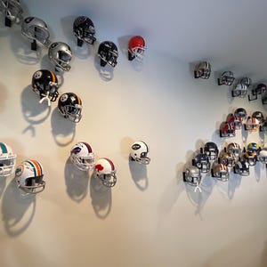 May include: A white wall with a collection of football helmets displayed on wall mounts. The helmets are in various colors and designs, including red, white, black, blue, and green.