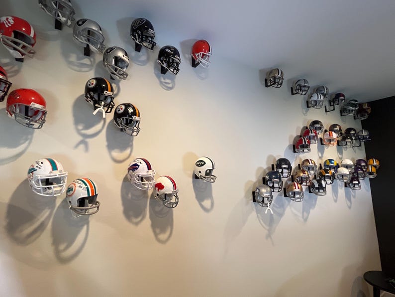 May include: A white wall with a collection of football helmets displayed on wall mounts. The helmets are in various colors and designs, including red, white, black, blue, and green.