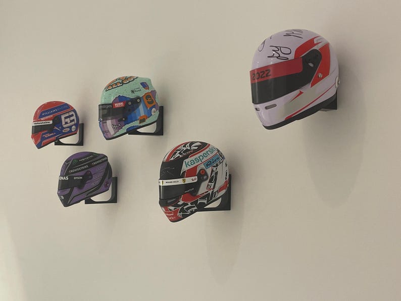 May include: Five racing helmets mounted on black wall mounts. The helmets are various colors and designs, including red, white, and blue, black and purple, and green, white, and blue. The helmets have sponsors' logos and names, including "Williams", "Kaspersky", and "222".