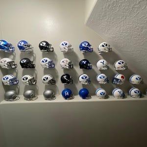 May include: A collection of miniature football helmets in various colors, including blue, white, and black. The helmets are arranged in rows on a white wall. Some helmets have the letter "Y" on them.