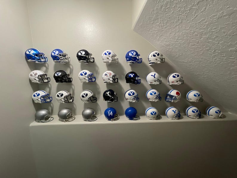 May include: A collection of miniature football helmets in various colors, including blue, white, and black. The helmets are arranged in rows on a white wall. Some helmets have the letter "Y" on them.