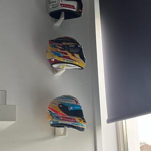 May include: Three racing helmets are displayed on a white wall. The helmets are white, yellow, and blue with various sponsors' logos, including Kaspersky, Santander, Epson, and Hublot.