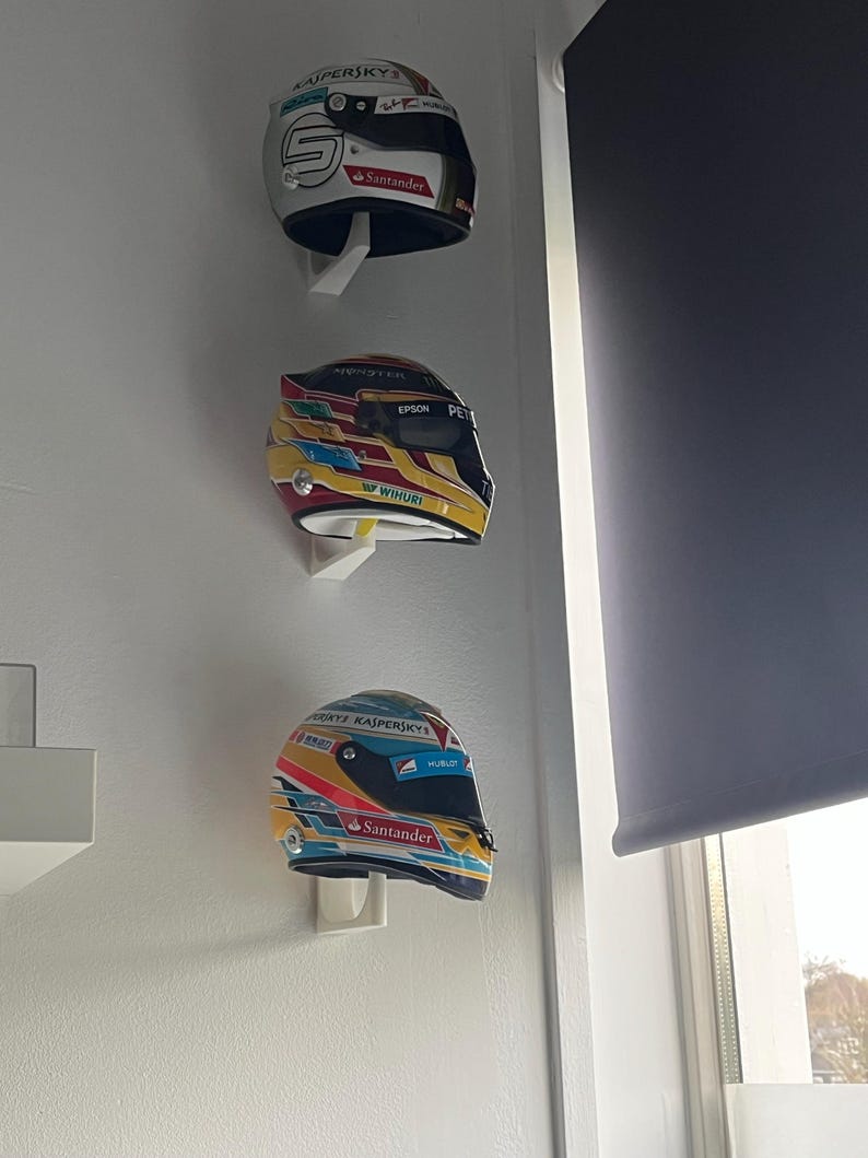 May include: Three racing helmets are displayed on a white wall. The helmets are white, yellow, and blue with various sponsors' logos, including Kaspersky, Santander, Epson, and Hublot.