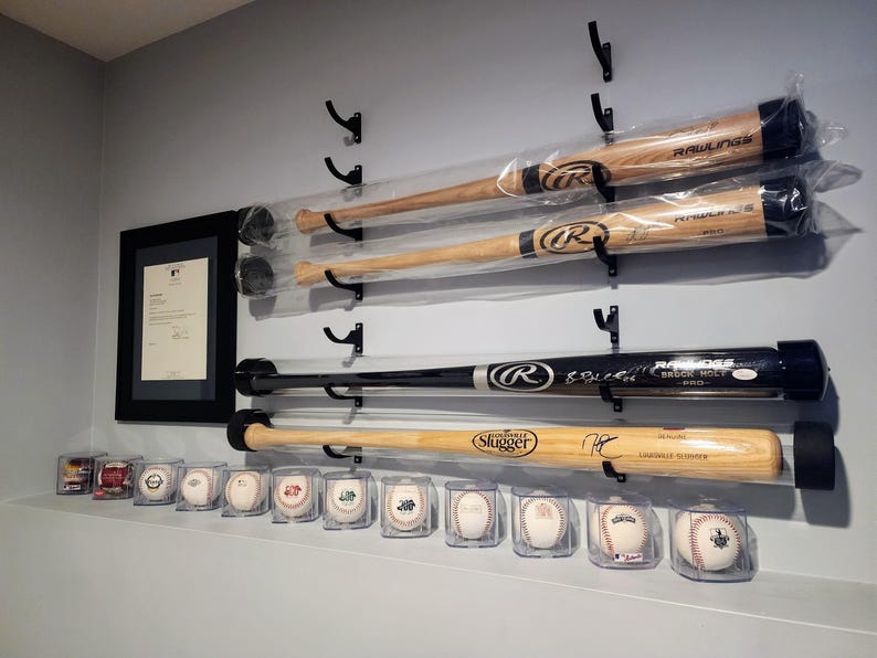 K&ouml;nnte beinhalten: Eine Wanddisplay mit Baseballschl&auml;gern und Baseballs. Die Schl&auml;ger befinden sich in durchsichtigen Plastikr&ouml;hren und sind mit schwarzen Haken an einer Wand befestigt. Die Baseballs befinden sich in durchsichtigen Plastikboxen und sind in einer Reihe auf einem Regal angeordnet. Die Schl&auml;ger sind von ber&uuml;hmten Baseballspielern signiert, darunter "Rawlings" und "Louisville Slugger".