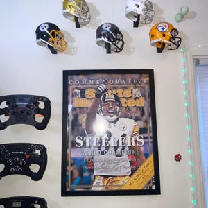 May include: A framed magazine cover featuring a football player in a black and yellow uniform with the text "Steelers World Champions" and "Super Bowl XL Commemorative".  The image also shows six football helmets in black, white, and gold with the Pittsburgh Steelers logo.